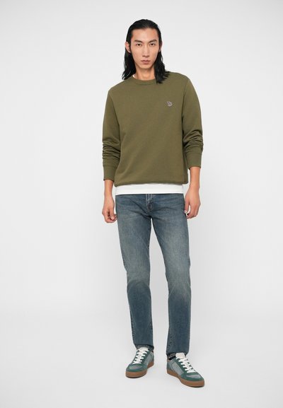 Sweatshirt vert olive avec des poignets côtelés, superposé à une chemise blanche, associé à un jean en denim bleu clair et des baskets teal avec des accents blancs.