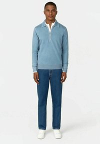 Ein Mann steht, trägt einen hellblauen Reißverschluss-Pullover über einem weißen Hemd, blaue Jeans und weiße Sneakers vor einem einfarbigen hellen Hintergrund.