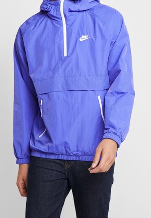 Veste coupe-vent en nylon violet avec un zip frontal mi-long, une capuche et des poignets élastiques. Elle possède deux poches latérales zippées blanches et un logo blanc sur la poitrine.