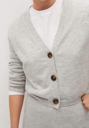 Jasnoszary dzianinowy kardigan z trzema brązowymi guzikami, noszony na białej koszuli, w zestawie z pasującymi jasnoszarymi spodniami.