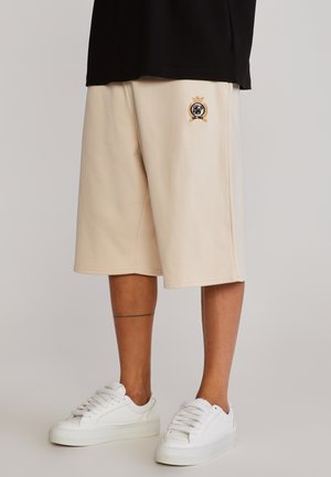 Bež šorc do koljena s izvezenim grbom, nošen uz bijele tenisice i crnu majicu vidljivu iznad šorca.