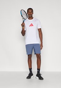 Biała koszulka Adidas z czerwonym logo, niebieskie spodenki i czarne tenisówki. Trzyma rakietę tenisową, stoi na jednolitym tle.