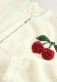 Fleece jacket in off-white with a zip closure. Features red plush cherry embroidery and green leaf accents. Soft texture, ribbed collar.