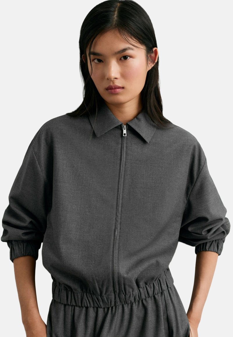 Jeune femme aux cheveux noirs et raides porte une veste zippée gris foncé avec poignets et taille élastiques sur un fond uni.
