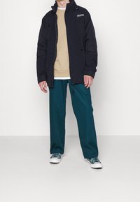 Svart isolerad jacka med hög krage, bärs över en beige sweatshirt, matchad med teal byxor och mönstrade sneakers.