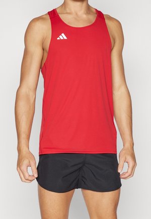 Mann trägt rotes ärmelloses Sportshirt mit weißem Logo und schwarze Laufshorts, steht vor einfarbigem Hintergrund.