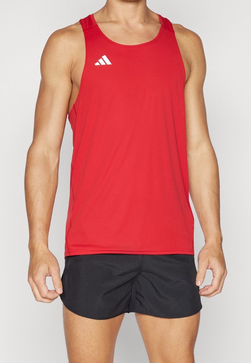 Homme portant un maillot de sport rouge sans manches avec un logo blanc et un short de course noir, debout devant un fond uni.
