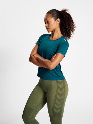 Kobieta z kręconymi włosami w morskim topie z krótkim rękawem i oliwkowych legginsach, stojąca z założonymi rękami, patrząca na bok na białym tle.