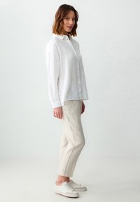 Camicia bianca in lino con maniche lunghe e chiusura a bottoni, abbinata a pantaloni in lino beige chiaro e sneakers bianche, in piedi contro uno sfondo semplice.