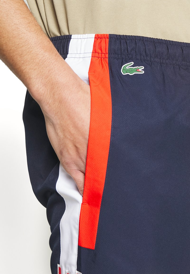 Shorts de sport bleu marine avec des bandes latérales orange et blanches, dotés d'une finition texturée et d'un logo Lacoste vert sur le côté gauche.