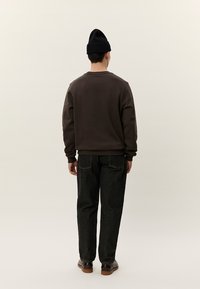 Hombre con gorro negro, sudadera marrón oscuro, vaqueros negros y zapatos marrones, de pie de espaldas contra un fondo beige liso.