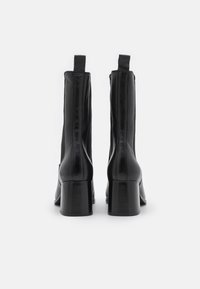 Bottes courtes en cuir noir à finition lisse, talons carrés et languettes à l'arrière. Présentent des coutures verticales sur les côtés.