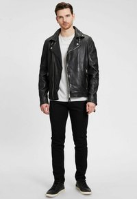 Schwarze Lederbikerjacke mit asymmetrischem Reißverschluss, Reverskragen und zwei Fronttaschen. Getragen über einem weißen Hemd und kombiniert mit schwarzen Jeans.