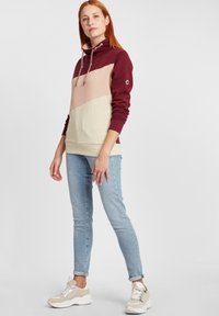 Fargeblokkert hettegenser med maroon, rosa og beige paneler. Den har en høy krage, snøring og ribbestrikkede mansjetter. Kombinert med lyseblå jeans og sneakers.