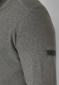 Grauer Strickpullover mit gerippten Akzenten an Schultern und Seiten sowie einem aufgenähten Logopatch am linken Ärmel.