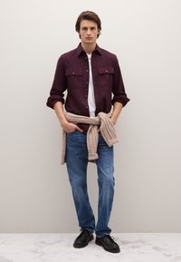 Bordeauxrotes Hemd mit Knöpfen und zwei Brusttaschen, kombiniert mit weißem T-Shirt, blauer Jeans und schwarzen Schuhen. Beiger Strickpullover, in der Taille gebunden.