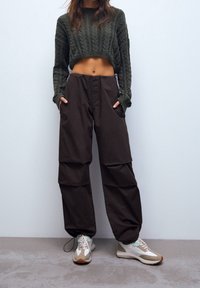 Donkerbruine, loszittende broek met zijzakken, gecombineerd met een cropped groene gebreide trui. De schoenen hebben een multikleurig ontwerp.