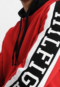 Rode hoodie met zwarte koorden en een zwart-wit omlijnd zwart stripe op de mouw met vetgedrukte witte tekst.