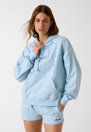 Femme portant un sweat à capuche clair à fleurs bleu clair et un short assorti avec le logo de la marque, debout devant un fond uni.
