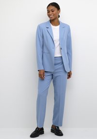 Světle modrý přiléhavý blazer a odpovídající kalhoty, vyrobené z hladké látky. Blazer má jeden knoflík a klopové límce; kalhoty mají elegantní střih.