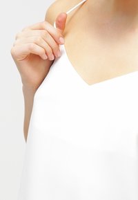 Vit linne med V-ringning och tunna axelband; slät, matt textur; hand placerad nära ringningen.