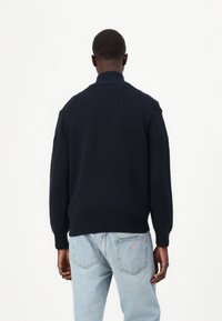 Marineblå strikkegenser med høy ribbekrage og senkede skuldre, sammen med lyseblå jeans med en trekantlogo på baksiden.