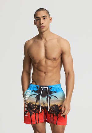 Uomo muscoloso in piedi con le mani in tasca, indossa pantaloncini da bagno colorati con stampa di palme e scritta "TERRANOVA" sul lato.
