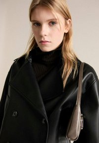 Manteau en cuir noir à double boutonnage avec larges revers, associé à un pull col roulé noir à côtes. Un petit sac marron pend d'une épaule.