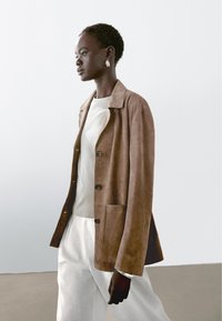 Braune Wildlederjacke mit Reverskragen, Knopfverschluss und Seitentaschen, kombiniert mit einem hellgrauen Hemd und weißen Hosen.