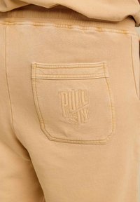 Pantalons de survêtement beiges avec une poche arrière portant le logo en relief "PULL IN". Tissu doux avec une taille élastique et des détails de couture visibles.