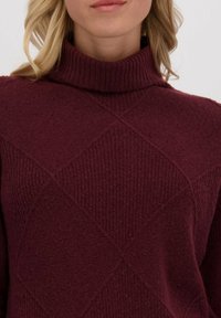 Vrouw in een bordeauxrode gebreide coltrui met een gestructureerd diamantpatroon, die de halslijn en bovenborst toont.