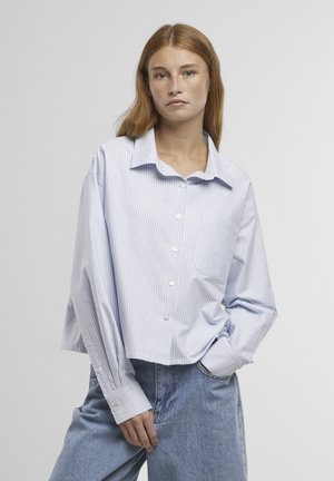 Mladá žena s dlhými červenými vlasmi v voľnej svetlomodrej pruhovanej košeli so zapínaním na gombíky a modrých džínsoch, stojaca s jednou rukou v vrecku.