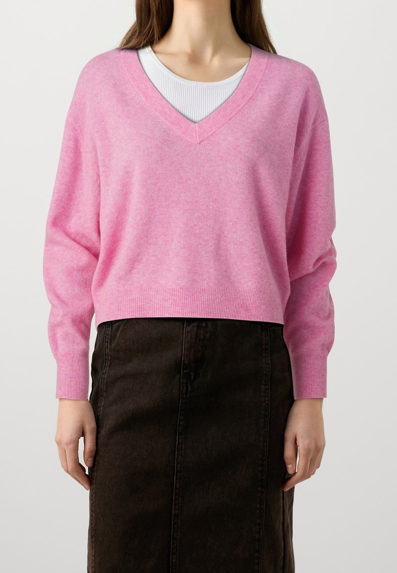 Kvinna iklädd en ljusrosa V-ringad tröja över en vit ribbad topp, kombinerad med en högmidjad svart denimkjol.