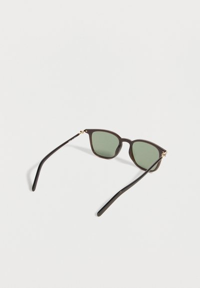 Γυαλιά ηλίου με σκούρα καφέ πλαίσια και πράσινους φακούς, ανοιχτά, ακουμπισμένα σε μια απλή λευκή επιφάνεια.