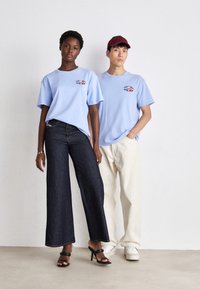 Dos modelos llevan camisetas de algodón de color azul claro con un logotipo impreso. Un modelo combina la camiseta con jeans anchos de color oscuro, mientras que el otro la lleva con pantalones color blanco roto.