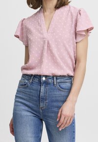 Różowa bluzka z krótkim rękawem i dekoltem w serek, z teksturowanym wzorem małych białych kwiatów. Połączona z klasycznymi niebieskimi dżinsami.