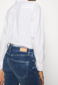 Weißes Hemd mit langen Ärmeln, kombiniert mit dunkelblauen, enganliegenden Jeans, die auf dem hinteren Bund ein Leder-Label haben.