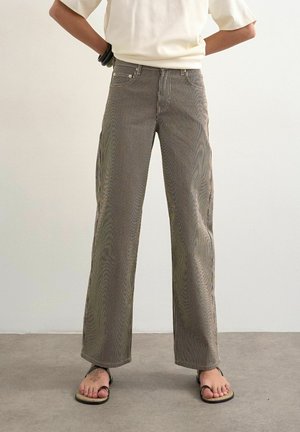 Personne portant un pantalon taille haute, jambes larges avec fines rayures verticales, un haut blanc, des bracelets noirs et des sandales, debout sur un sol gris.