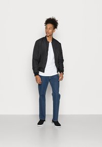 Chaqueta bomber negra con puños acanalados, camiseta blanca, jeans azules y zapatillas negras con suelas blancas; de pie sobre un fondo liso.