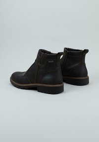 Par de botas de cuero negro hasta el tobillo con costuras marrones, cremallera lateral, tira de agarre y suelas de goma gruesas sobre un fondo liso gris claro.
