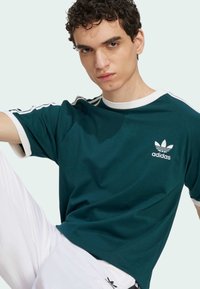 T-shirt en coton bleu foncé avec un col et des accents de manches blancs. Présente un petit logo Adidas blanc sur la poitrine. Pantalons blancs partiellement visibles.