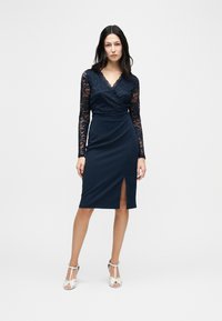 Γυναίκα φορούσα μπλε σκούρο μπλούζα από δαντέλα με μακριά μανίκια και εφαρμοστή φούστα με σκίσιμο μπροστά, συνδυασμένα με λευκές πλατφόρμες τακουνιών, στέκεται μπροστά σε λευκό φόντο.