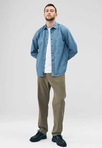 Een man staat zelfverzekerd, gekleed in een lichtblauw overhemd over een wit T-shirt, met olijfgroene broek en zwarte schoenen, voor een grijze achtergrond.
