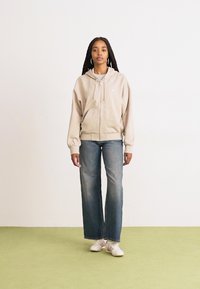 Beige hettegenser med glidelås, snor, frontlommer og ribbede mansjetter, sammen med falmede blå vide bukser og hvite sneakers.