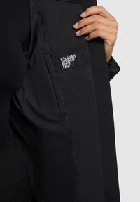 Interno di un piumino nero con una texture liscia, dotato di un'etichetta del marchio visibile con scritte bianche e un particolare con tasca con zip.