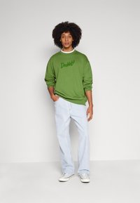 Felpa verde con logo "Doubtof" ricamato, vestibilità ampia, abbinata a jeans a gamba larga blu chiaro e sneakers bianche. Sfondo semplice.