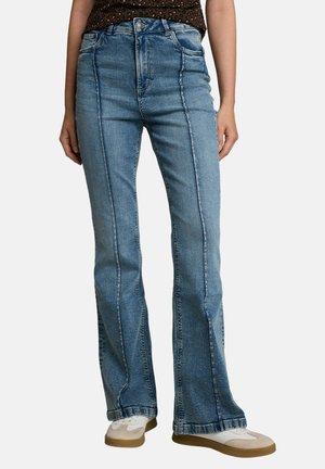 Frontansicht einer Person, die blaue hoch taillierte Jeans mit sichtbaren vertikalen Nähten und weiße Sneakers mit beigen Sohlen trägt.