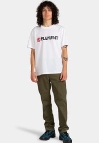 Biała koszulka z krótkim rękawem z czarnym napisem "ELEMENT" i czerwonym logo, w połączeniu z oliwkowym zielonymi spodniami. Model nosi niebiesko-czarne buty.