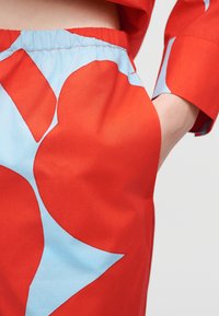 Κοντινό πλάνο φούστας με κόκκινο και ανοιχτό μπλε μοτίβο, με ελαστική μέση και χέρι στην τσέπη, μπροστά σε απλό φόντο.