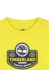 T-shirt en coton jaune avec un col rond, des boutons-pression sur l'épaule, et un logo graphique noir représentant un arbre et du texte.